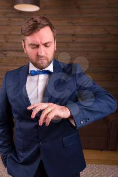 Portrait of groom in suit and bow-tie looking on a wedding ring on his finger, wooden room interior on background