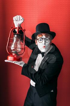 Pantomime actor performing with kerosene lantern. Comedy mime artist in suit, gloves, glasses, make-up mask and hat. Comedian male person with vintage oil lamp