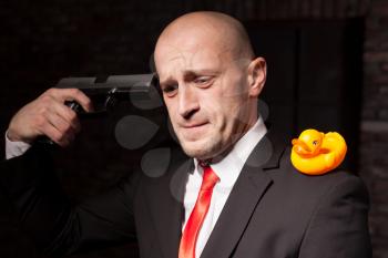 Contract murderer in suit and red tie with toy duck on shoulder aims a pistol in his head. Professional secret agent nervous breakdown concept