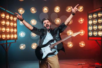 Male rock star with electro guitar hands up on the stage with the decorations of lights. Music entertainment