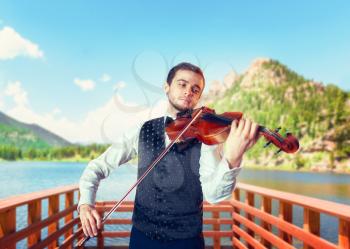 Male fiddler playing classical music on violin on wooden pier, lake and mountains on background. Violinist man with musical instrument plays on nature