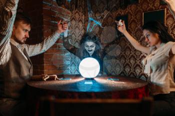Female foreteller calls the spirits, witchcraft. Scary witch reads a magic spell over a crystal ball, young people hands up on spiritual seance