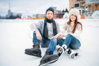 Young love couple in skates sitting on ice, skating rink. Winter ice-skating on open air, active leisure