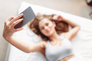 Attractive woman in underwear lies in bed and makes selfie on phone. Girl wake up in the morning in bedroom