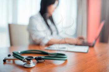 Stethoscope on wooden table closeup, family doctor concept, specialist works on laptop on background. Professional health care