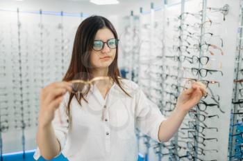 Young female buyer holds many glasses in hand, optics store, showcase with spectacles on background. Professional eyecare in glasses shop concept, eyeglasses choice
