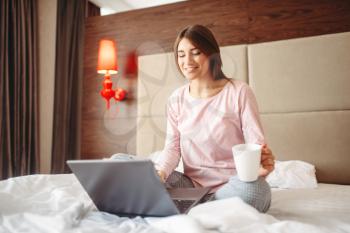 Female person in pajama drinks coffee and uses laptop in bed, good morning, bedroom interoir on background