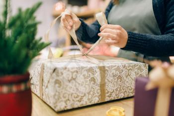 Female person ties a gold bow on gift box, handmade wrapping and decoration process. Woman wraps present on the table, decor procedure