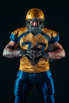 Muscular american football player in uniform and helmet holds ball in hands, black background