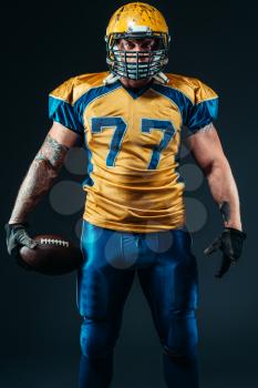 Muscular american football player in uniform and helmet holds ball in hands, black background