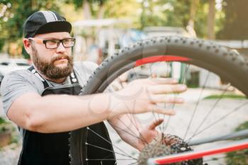 Bicycle repairman works with bike wheel, cycle workshop outdoor. Bearded mechanic in apron