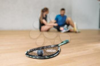 Squash game concept, racket with ball, young couple sitting on the floor after training, indoor sport club