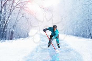 Ice hockey player in uniform on frozen walkway, winter forest on background. Ice-skating outdoors. Cold season sport