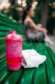 Sport bottle and towel on a bench in summer park, woman in sportswear relax on background
