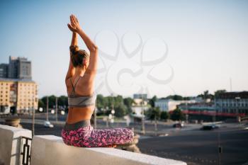Young woman sitting in yoga pose, city on background. Yogi training outdoors, relaxation exercise