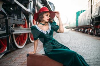 Young old-fashioned woman sitting on suitcase against vintage steam train, red wheels closeup. Old locomotive. Railway engine, travel by railroad