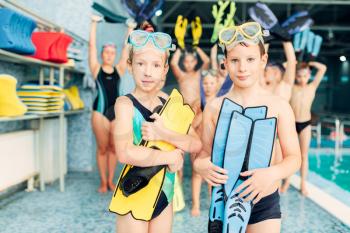 Portrait of boy and girl with colorful flippers in hands near swimming pool. Sportive healthy children after swimming exercises. Happy kids in modern sport center.