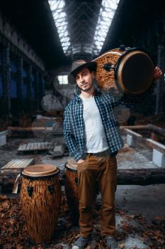 Young male drummer holds wooden drum on the shoulder, factory shop on background. Djembe, musical percussion instrument, 