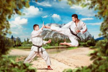 Martial arts karate masters in white kimono and black belts, outdoors practice, kick in jump, nature and mountains on background