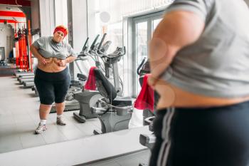 Overweight sweaty woman watches on her body against mirror in gym. Calories burning, obese female person in sport club