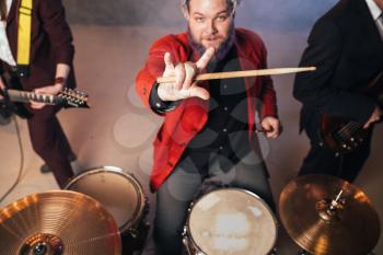 Drummer in red suit, performing on the stage of night club, electro and bas-guitarists on background. Rock band concert, music show