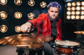 Bearded drummer in red suit on the stage with lights, retro style. Musical performer with colorful hair, drum instrument
