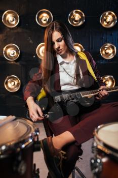 Female rock guitarist in suit sitting behind the drum kit, stage with lights on background, retro style. Live music concert