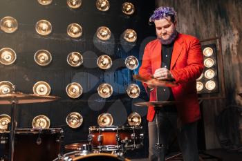 Bearded drummer in red suit on the stage with lights, retro style. Musical performer with colorful hair
