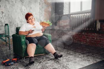 Overweight woman with sandwich in hands sits in a chair and watches TV, bulimic, fatty. Unhealthy lifestyle, obesity
