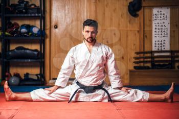 Martial arts karate master in white kimono and black belt doing stretching exercise in gym