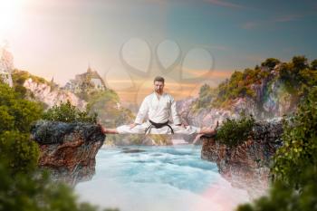 Martial arts master in white kimonodo sitting on the splits, stretching exercise over beautiful lake, mountains on background, outdoor training