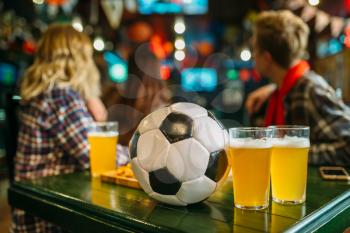 Ball and beer on the table in sports bar, football fans on background. Tv broadcasting, watching the game concept