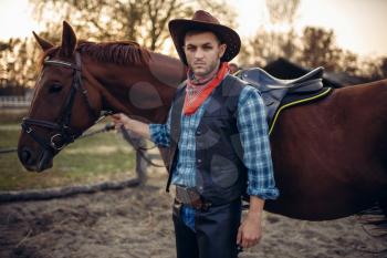 Brutal cowboy in jeans and leather jacket poses with horse on texas ranch, western. Vintage male person with animal, wild west