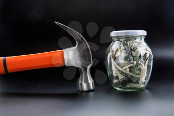 Money saving control concept, hammer against glass jar full of dollars. Cash economy