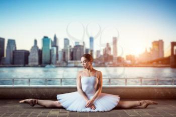 Body flexibility of classical ballet dancer. Ballerina in white dress sits on a twine, front view, cityscape with skyscrapers on background