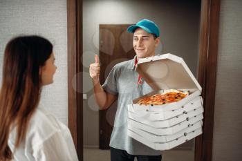 Cheerful pizza delivery boy shows thumbs up symbol to female customer, fast delivering service. Courier from pizzeria holds cardboard packages indoors, speed deliver