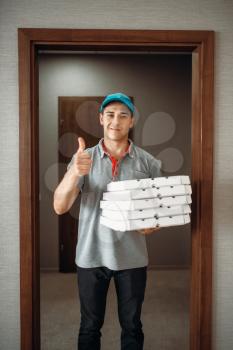 Cheerful pizza delivery boy shows thumb up symbol, delivering service. Courier from pizzeria holds cardboard packages indoors