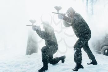 Two paintball players in uniform and masks shooting at the enemy, side view, winter forest battle.