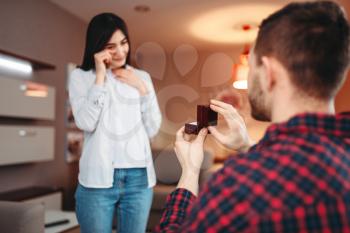 Young man makes an offer to get married to surprised woman and gives a ring. Modern apartment interior on background. Love couple relationship