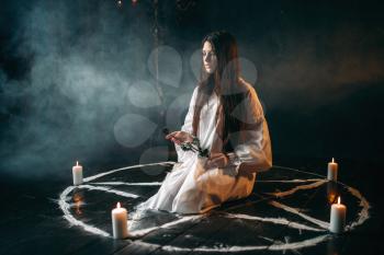 Young woman in white shirt holds burnt rose in hands, pentagram circle with candles, smoke all around. Dark magic ritual, occultism