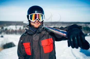 Skier in helmet and glasses holds skis and poles in hands, blue sky and snowy mountains on background. Winter active sport, extreme lifestyle. Downhill skiing