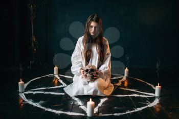 Young woman in white shirt holds human skull in hand, pentagram circle with candles, smoke all around, witch. Dark magic ritual, occult