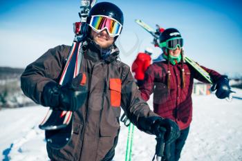Male and female skiers poses with skis and poles in hands, blue sky and snowy mountains on background. Winter active sport, extreme lifestyle. Downhill skiing