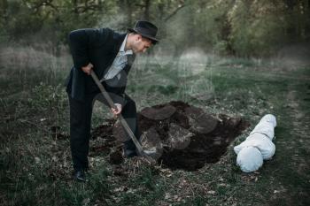 Male murderer with a shovel is digging a grave for the victim, the body wrapped in a canvas, serial maniac concept