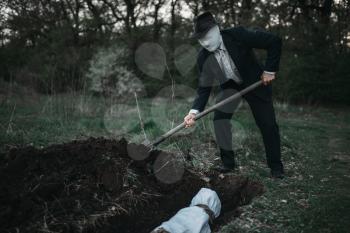 Bloody murderer is digging a grave for the victim in forest, the body wrapped in a canvas, serial maniac concept