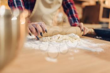 Housewife in an apron rolls out the dough with a rolling pin, kitchen interior on background. Female cook prepares fresh homemade cake. Domestic pie preparation