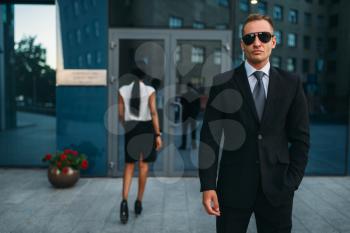 Serious bodyguard in suit and sunglasses, black business woman on background. Security guard is a risky profession, protection of VIPs, guarding