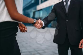Business woman and businessman shake hands, outdoors meeting of partners, modern office building on background, partnership negotiations. Successful businesspeople