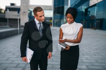 Business woman with phone and businessman with laptop in hands walking outdoors, modern office building on background, partnership negotiations during the lunch break. Successful businesspeople