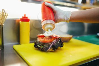 Male cook puts tomatoes in to the burger, fast food cooking. Hamburger preparation process, fastfood